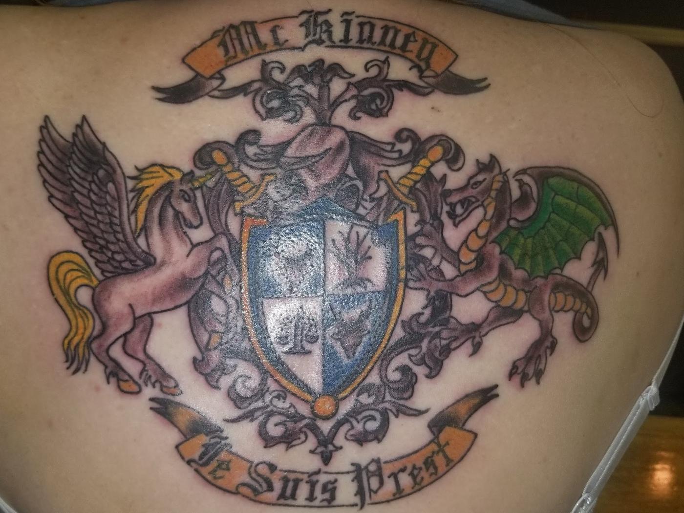 A large ornate crest tattoo with wings and a banner, across a back